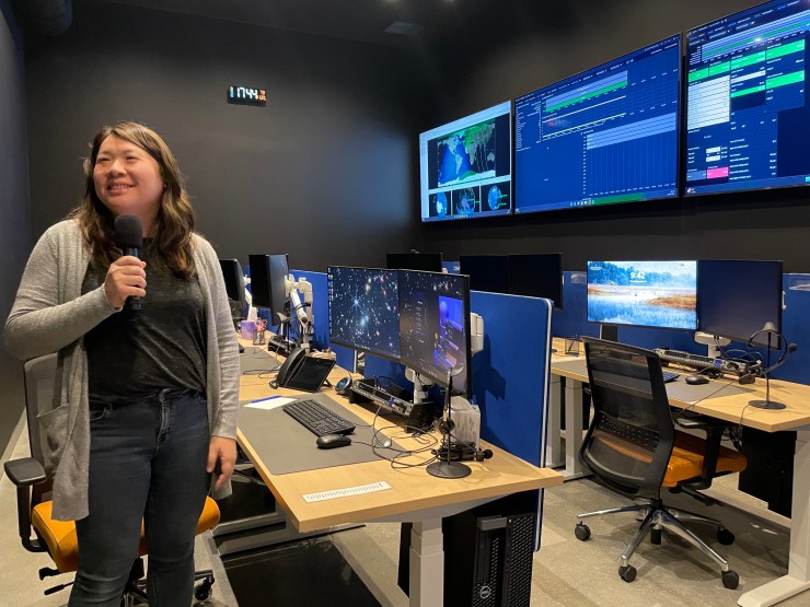 Wendy Shimata holds a microphone and stands in a room with screens on the wall and multiple computers.
